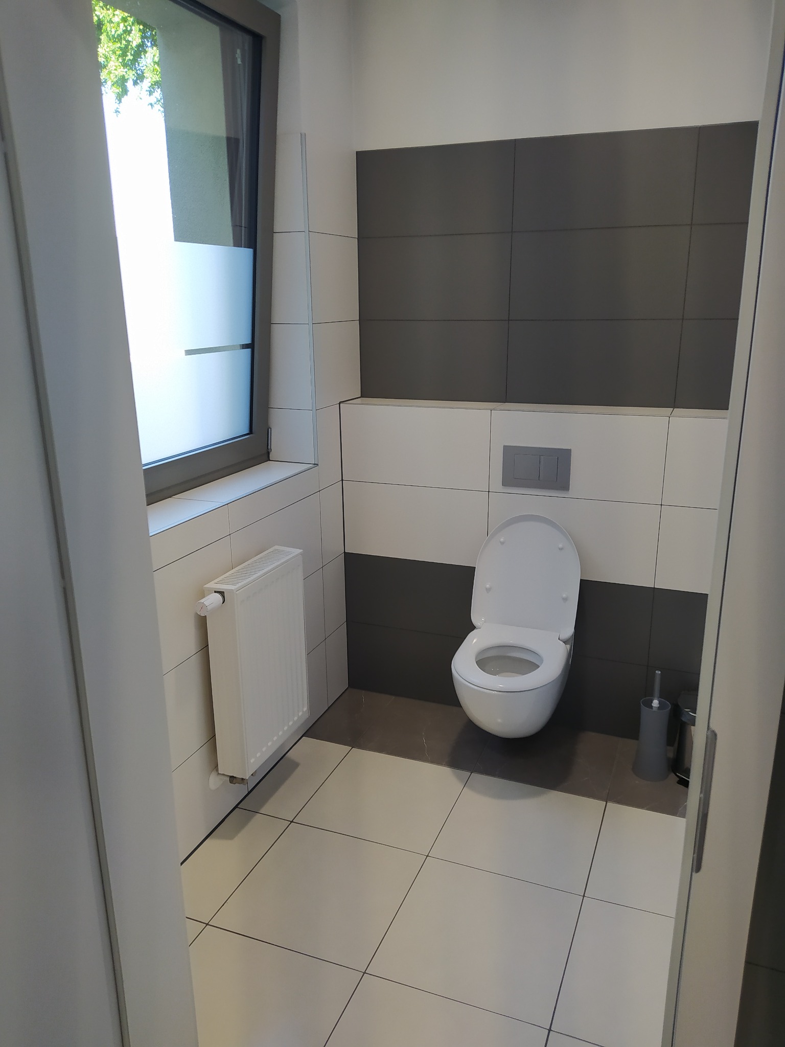 Nowoczesna łazienka w odcieniach bieli i szarości z podwieszaną toaletą, grzejnikiem drabinkowym pod oknem z częściowo zamrożoną szybą i dużymi kwadratowymi płytkami na podłodze i ścianach.