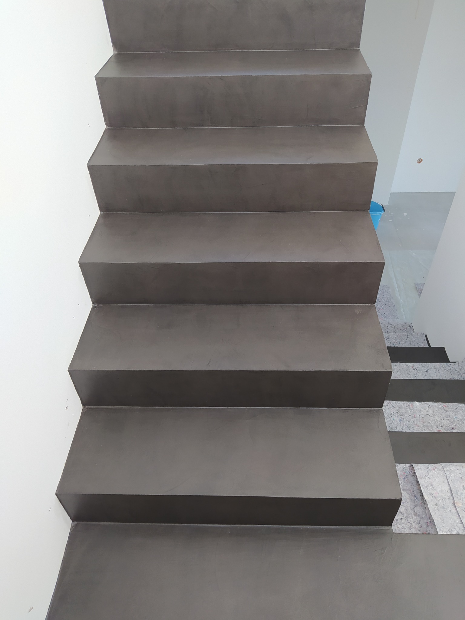 Schody betonowe w odcieniach szarości, widok z góry, z widocznym fragmentem białej ściany po lewej stronie i zabezpieczeniem podłogi na dole schodów.