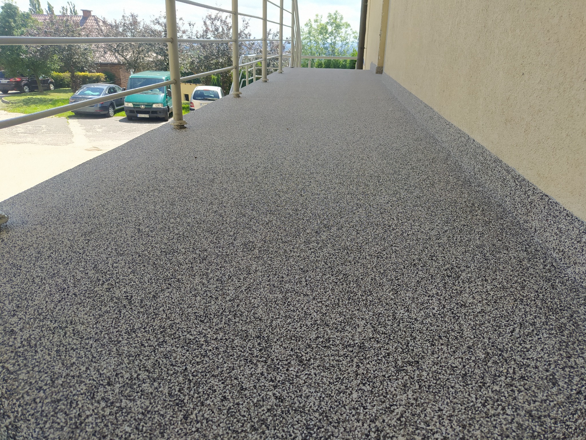 Szara, drobnoziarnista posadzka żywiczna na balkonie z metalową balustradą, widok z niskiej perspektywy, w tle zaparkowane samochody i zieleń.