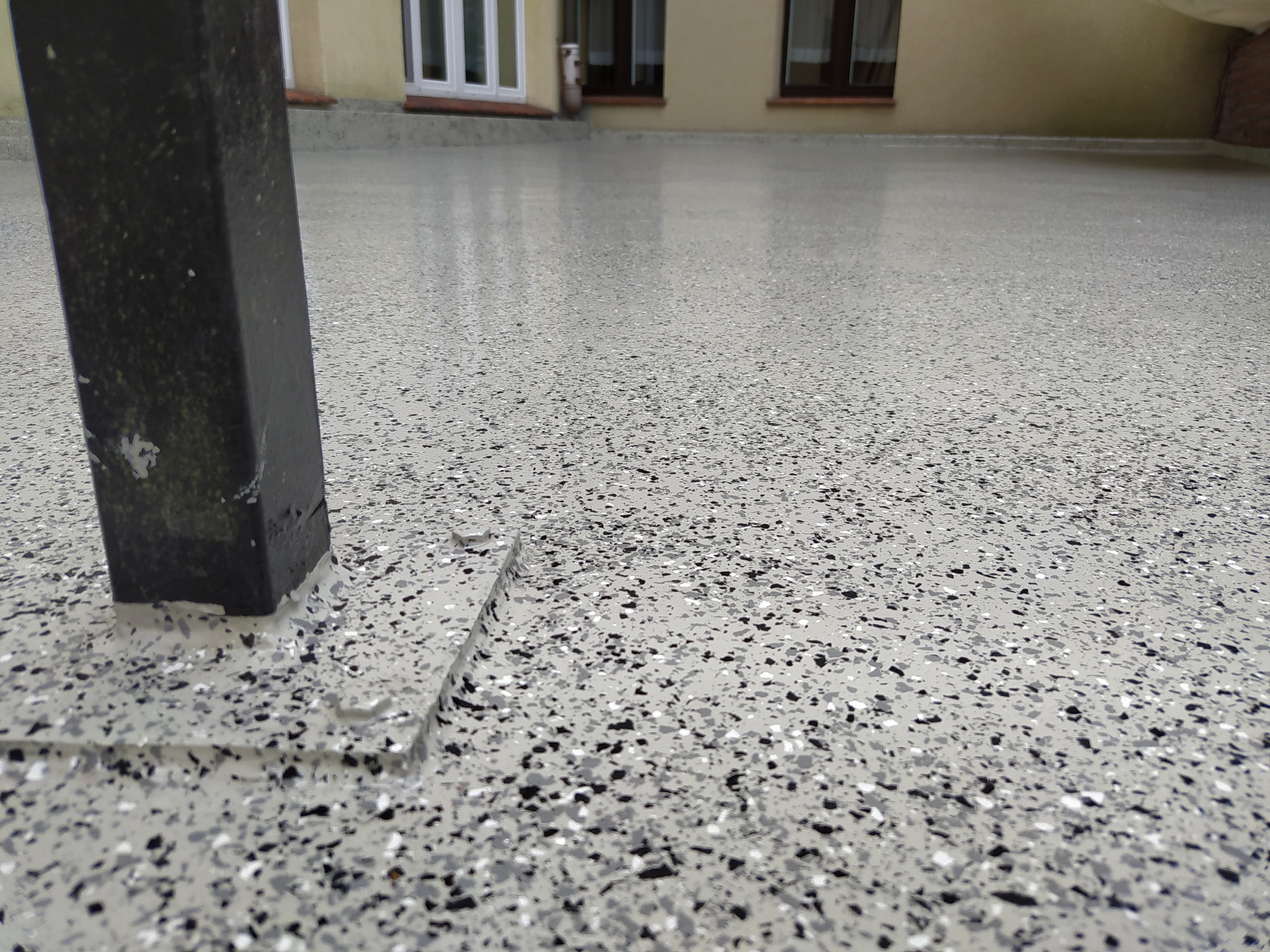 Szara, błyszcząca posadzka żywiczna z czarnymi i białymi wtrąceniami, otaczająca podstawę czarnego słupka, z widocznym profilem dylatacyjnym i fragmentem elewacji budynku w tle.