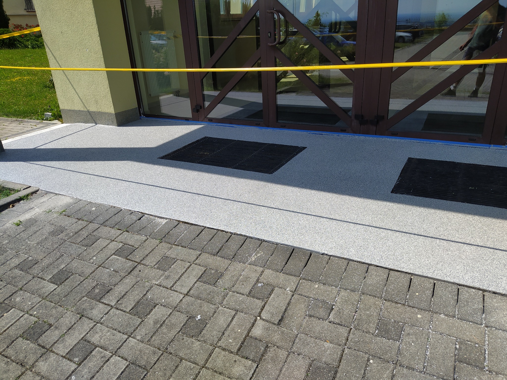 Nowa, jasnoszara posadzka żywiczna przed wejściem do budynku, zabezpieczona żółtą taśmą ostrzegawczą, z widocznymi kratkami wentylacyjnymi i fragmentem chodnika z kostki brukowej.