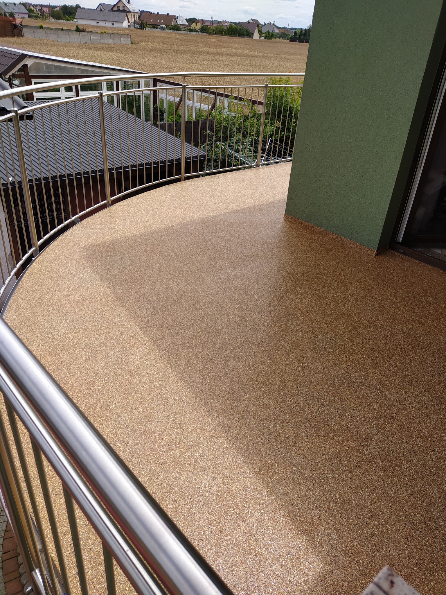 Beżowa, żywiczna posadzka na balkonie z metalową balustradą, widok na okoliczne domy i pole w tle.