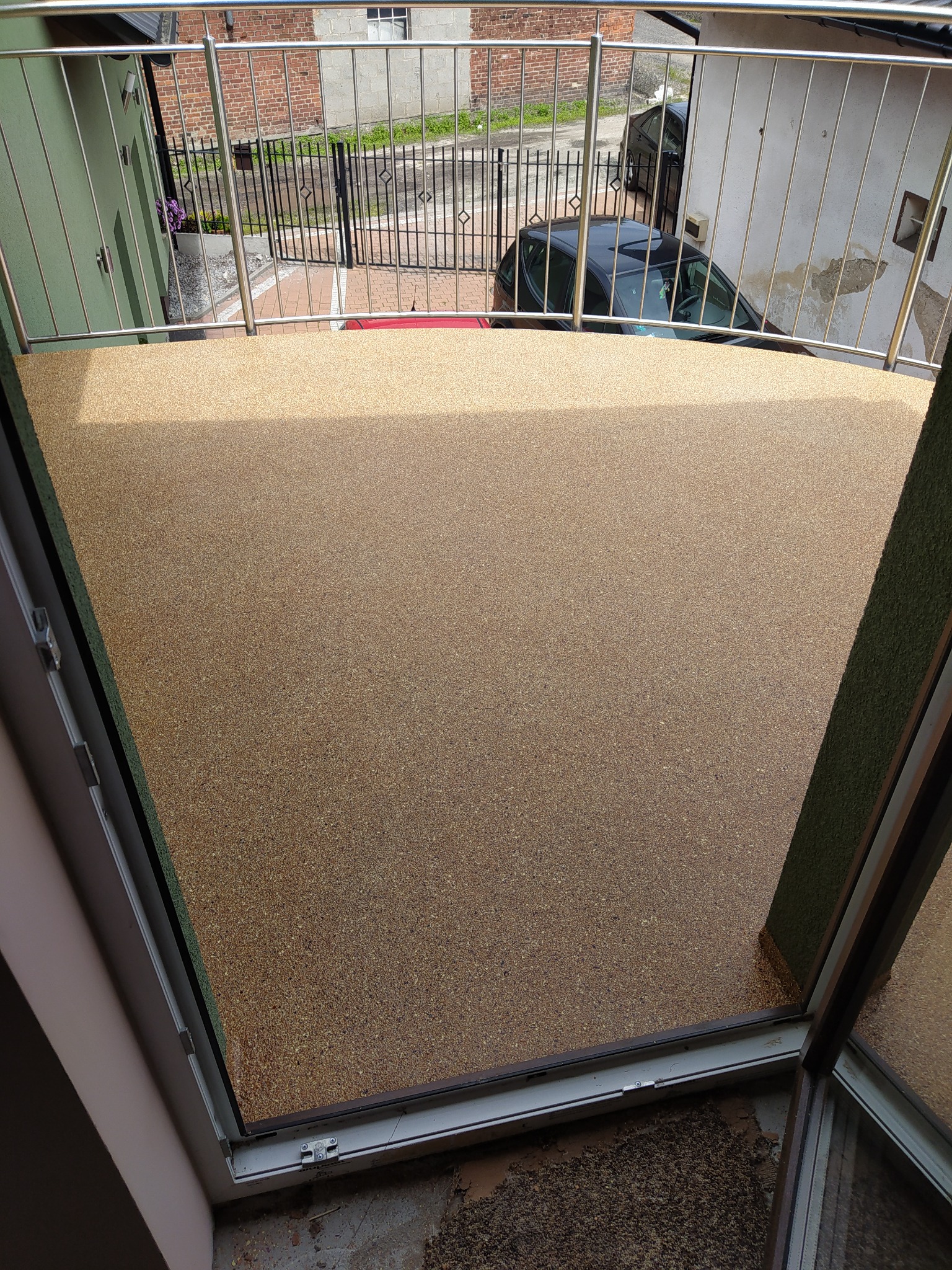 Balkon z nową, żywiczną posadzką w odcieniach beżu i brązu, widok z góry przez otwarte drzwi balkonowe, widoczne metalowe barierki balkonu i elementy otoczenia budynku.