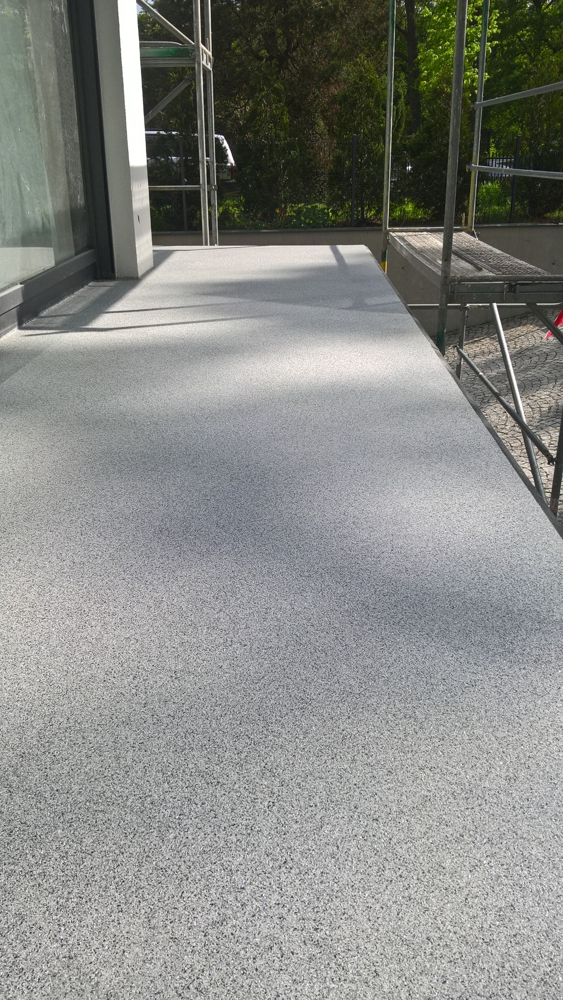 Szara, teksturowana posadzka balkonu z widocznymi rusztowaniami w tle, okno przesuwne z lewej strony kadru.