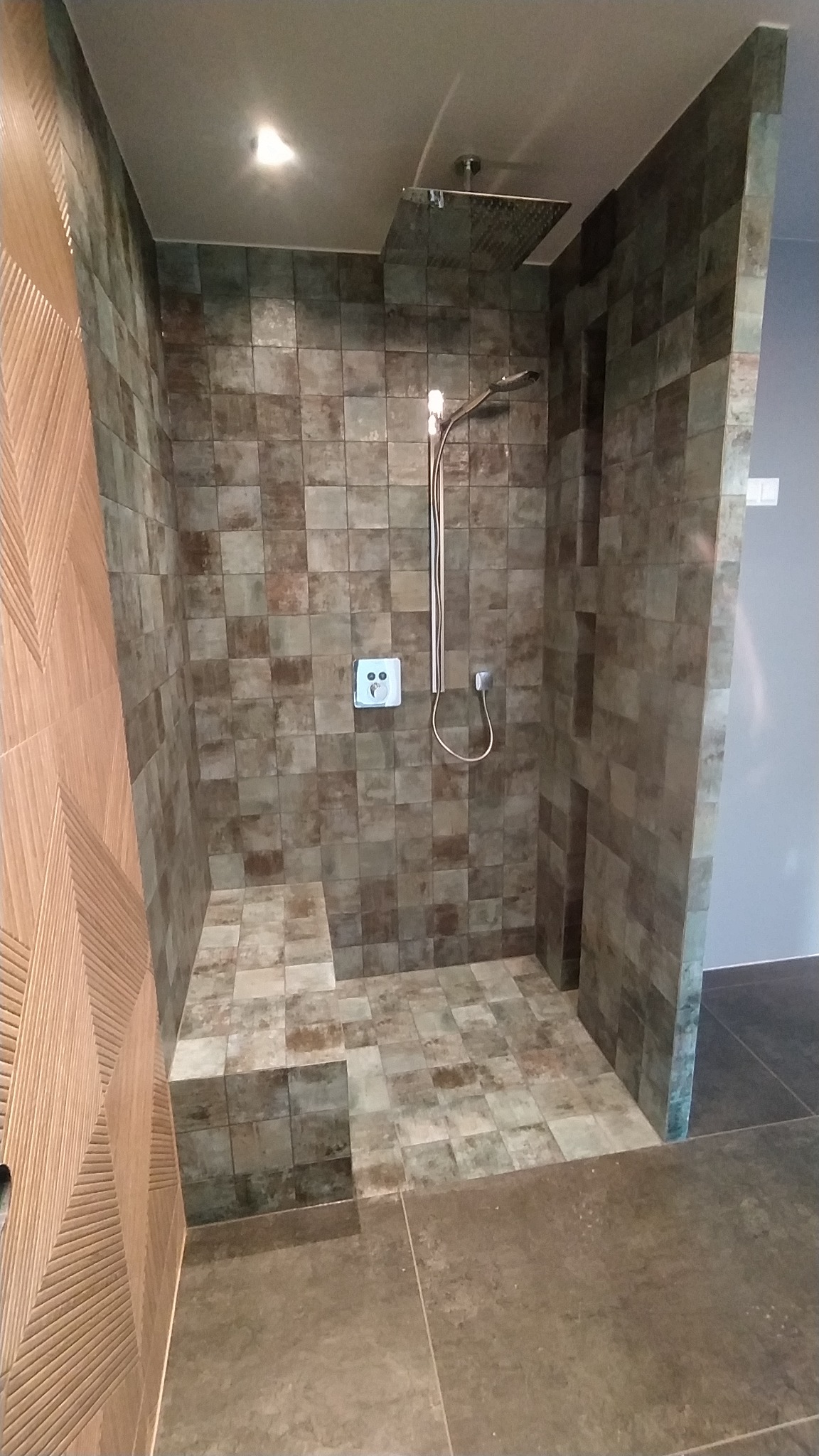 Nowoczesna kabina prysznicowa wyłożona kwadratowymi, brązowo-szarymi płytkami imitującymi kamień, z deszczownicą i siedziskiem, oświetlona punktowym światłem.