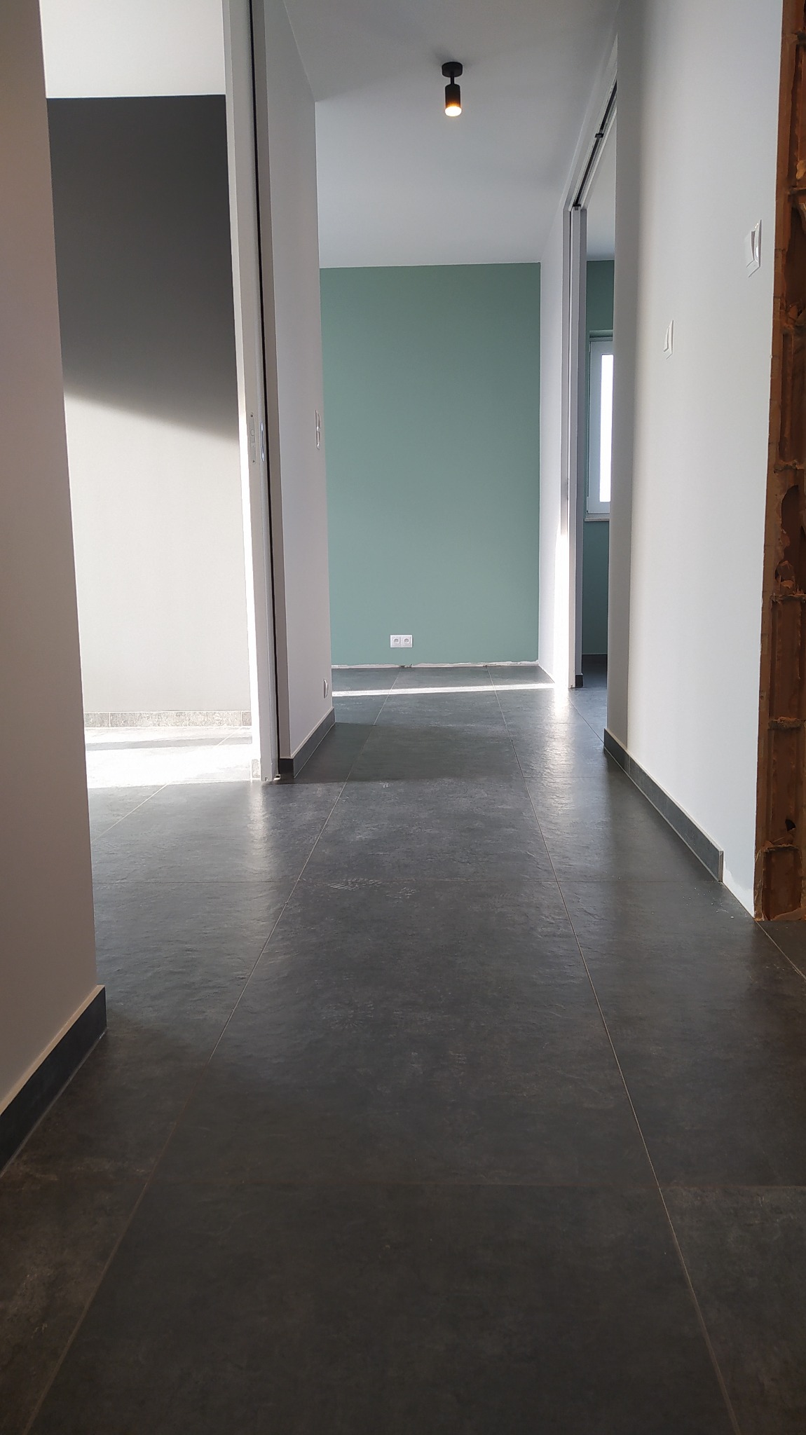 Korytarz z ciemną, matową podłogą wyłożoną płytkami, świeżo pomalowane ściany w odcieniach szarości i zieleni, widoczne fragmenty surowej ściany z cegły po prawej stronie.