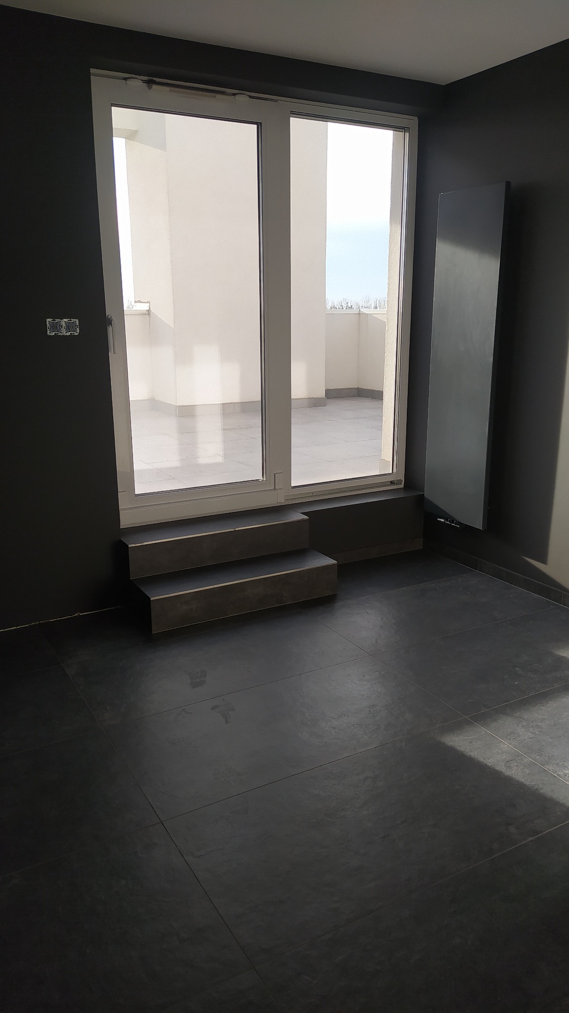 Nowoczesne wnętrze z ciemną podłogą z płytek, dwuskrzydłowymi drzwiami balkonowymi z niskim progiem i schodkami w kolorze podłogi oraz grzejnikiem panelowym na ścianie.