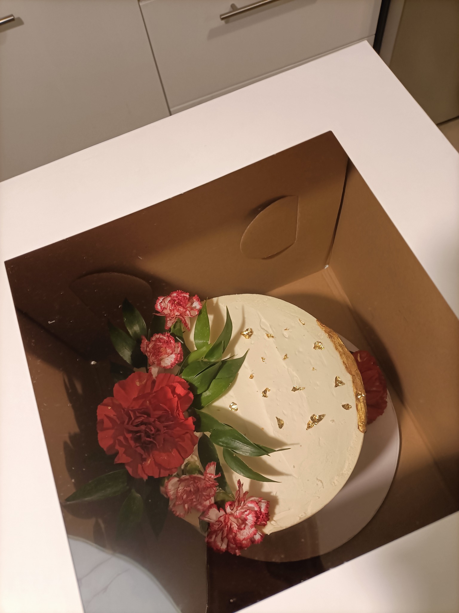 Tort z kremem, dekorowany czerwonymi goździkami i zielonymi liśćmi, posypany płatkami złota, umieszczony w kartonowym pudełku.