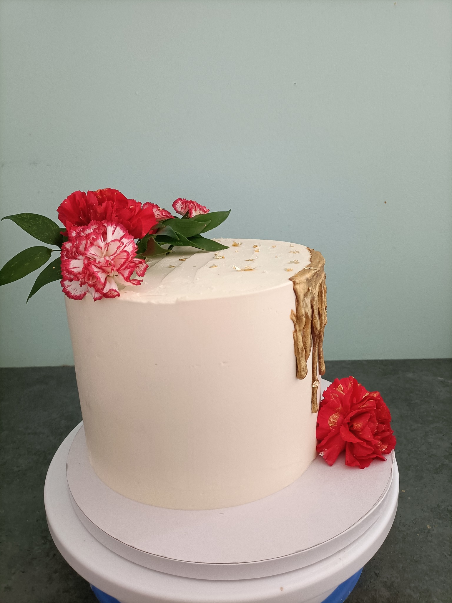 Tort z białym kremem, złotymi dripami i dekoracją z czerwonych goździków oraz złotych płatków, umieszczony na obrotowym stojaku.