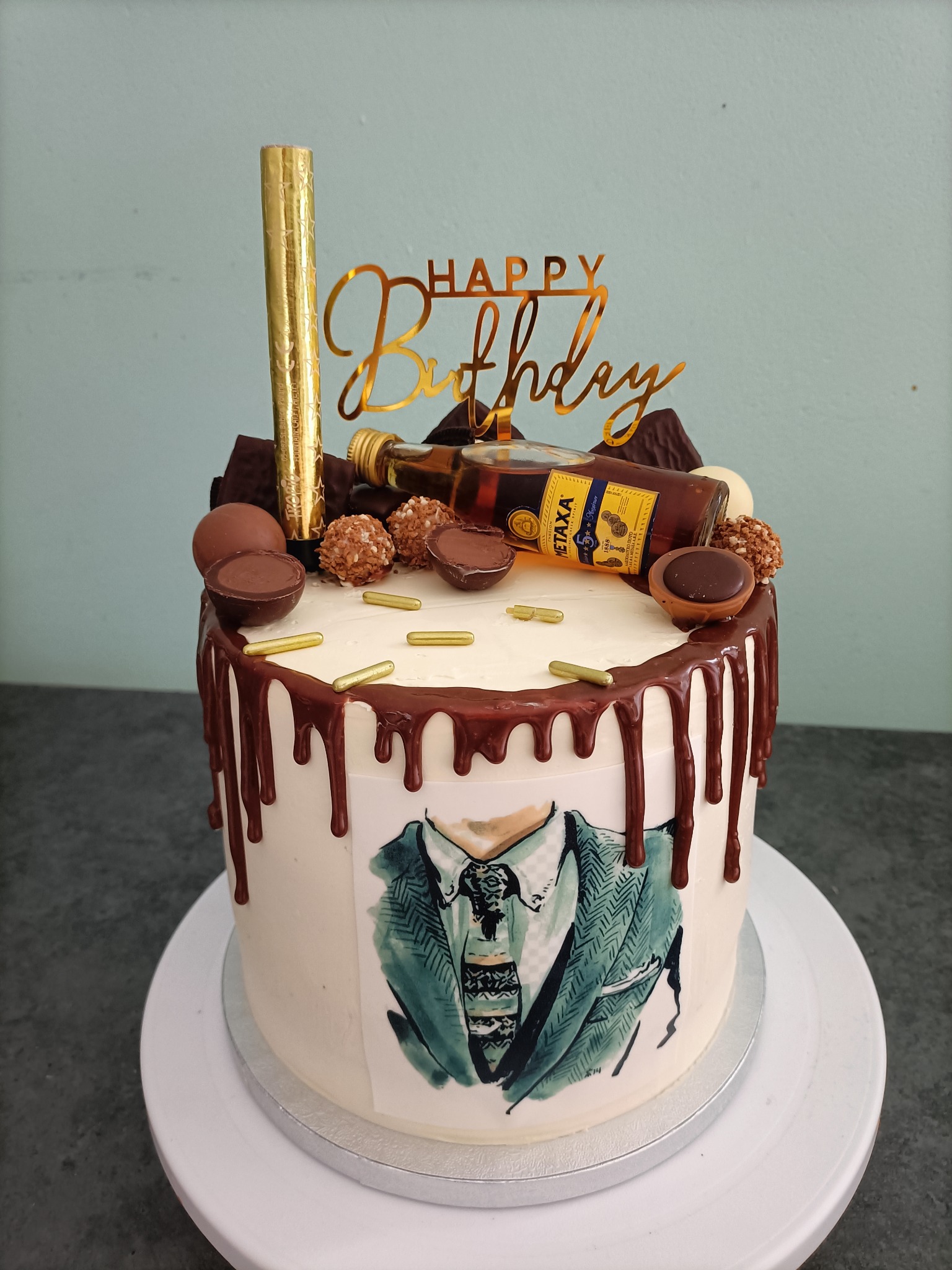 Tort urodzinowy dekorowany czekoladą, pralinkami, złotym topperem 'Happy Birthday', małą butelką alkoholu i rysunkiem marynarki w stylu akwareli.