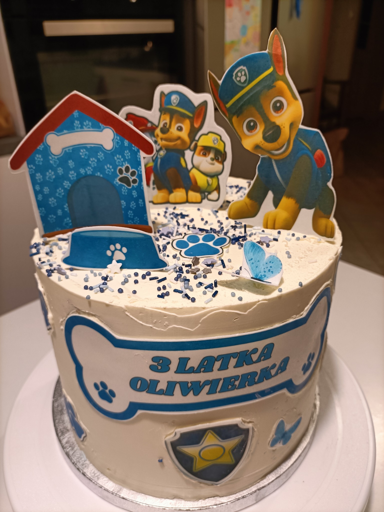 Tort urodzinowy dla dziecka ozdobiony postaciami z bajki 'Psi Patrol', z napisem '3 Latka Oliwiera' na białym kremie, posypany niebieskimi i białymi kuleczkami.