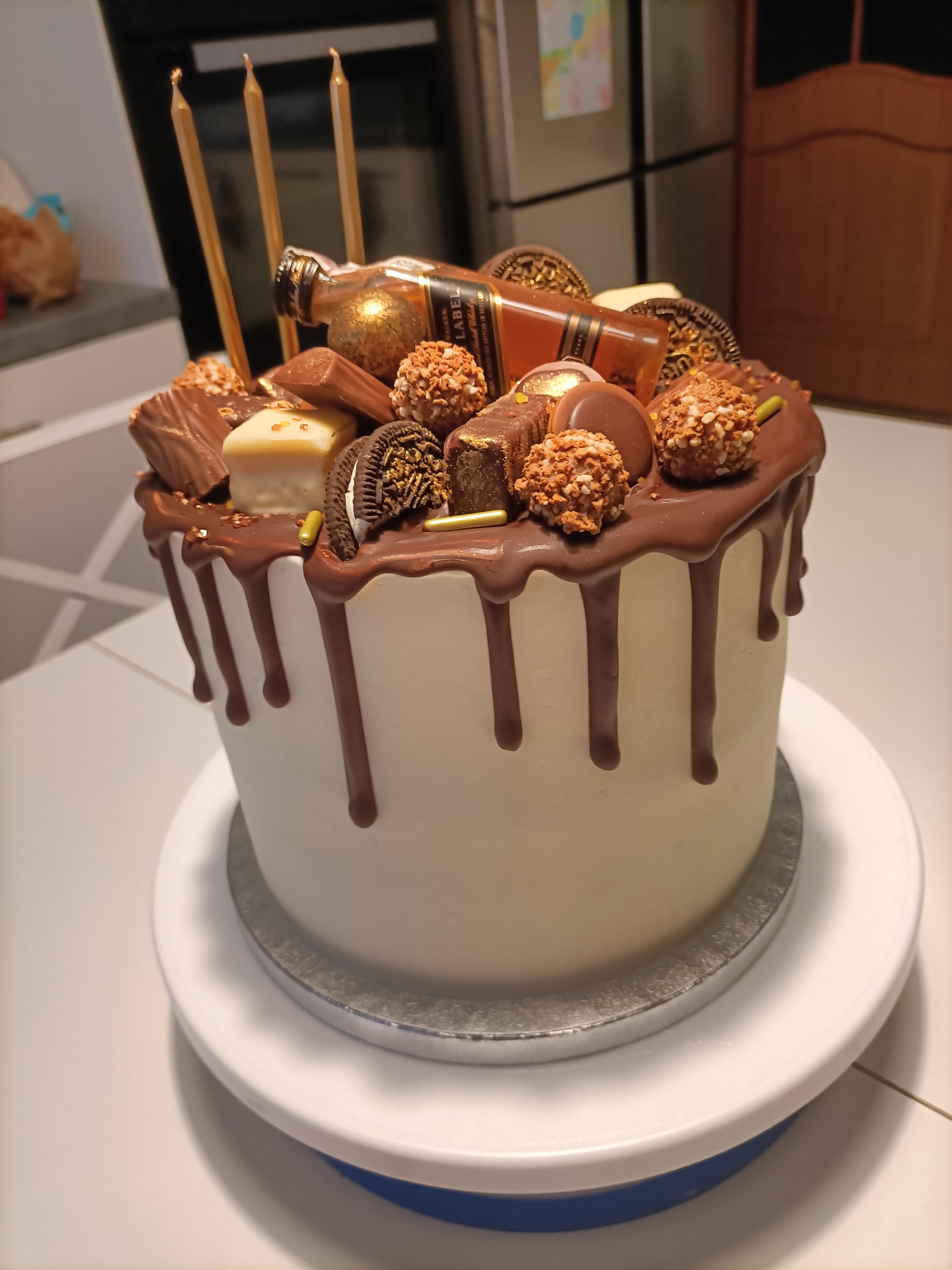 Tort urodzinowy z polewą czekoladową, dekorowany batonikami, ciasteczkami Oreo, złotymi kulkami i miniaturową butelką alkoholu leżącą na boku, całość na obrotowej podstawie.