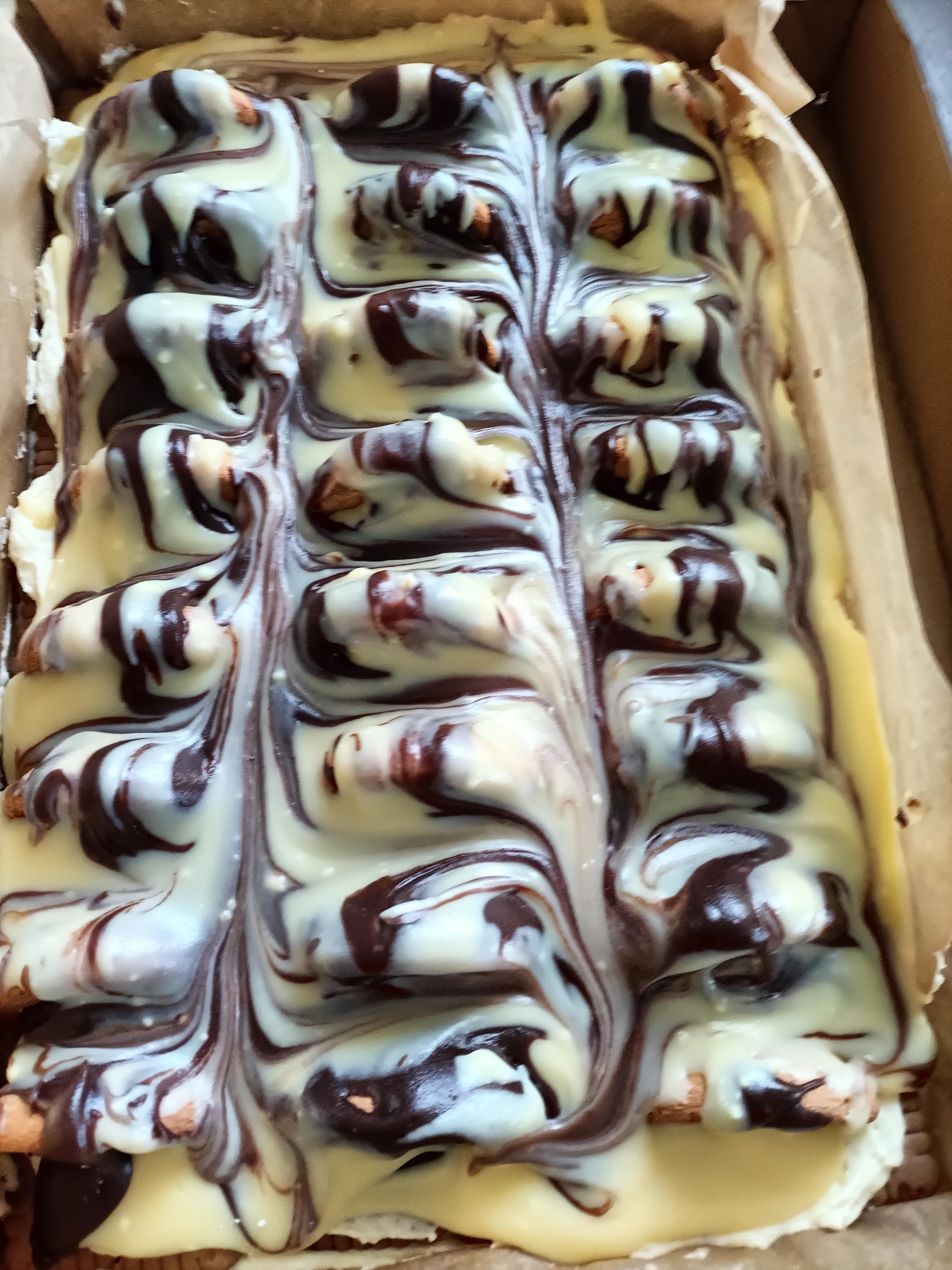 Kremowy blok ciasta z polewą z ciemnej i białej czekolady, widoczne fragmenty ciasta i orzechów, całość w papierowej foremce.