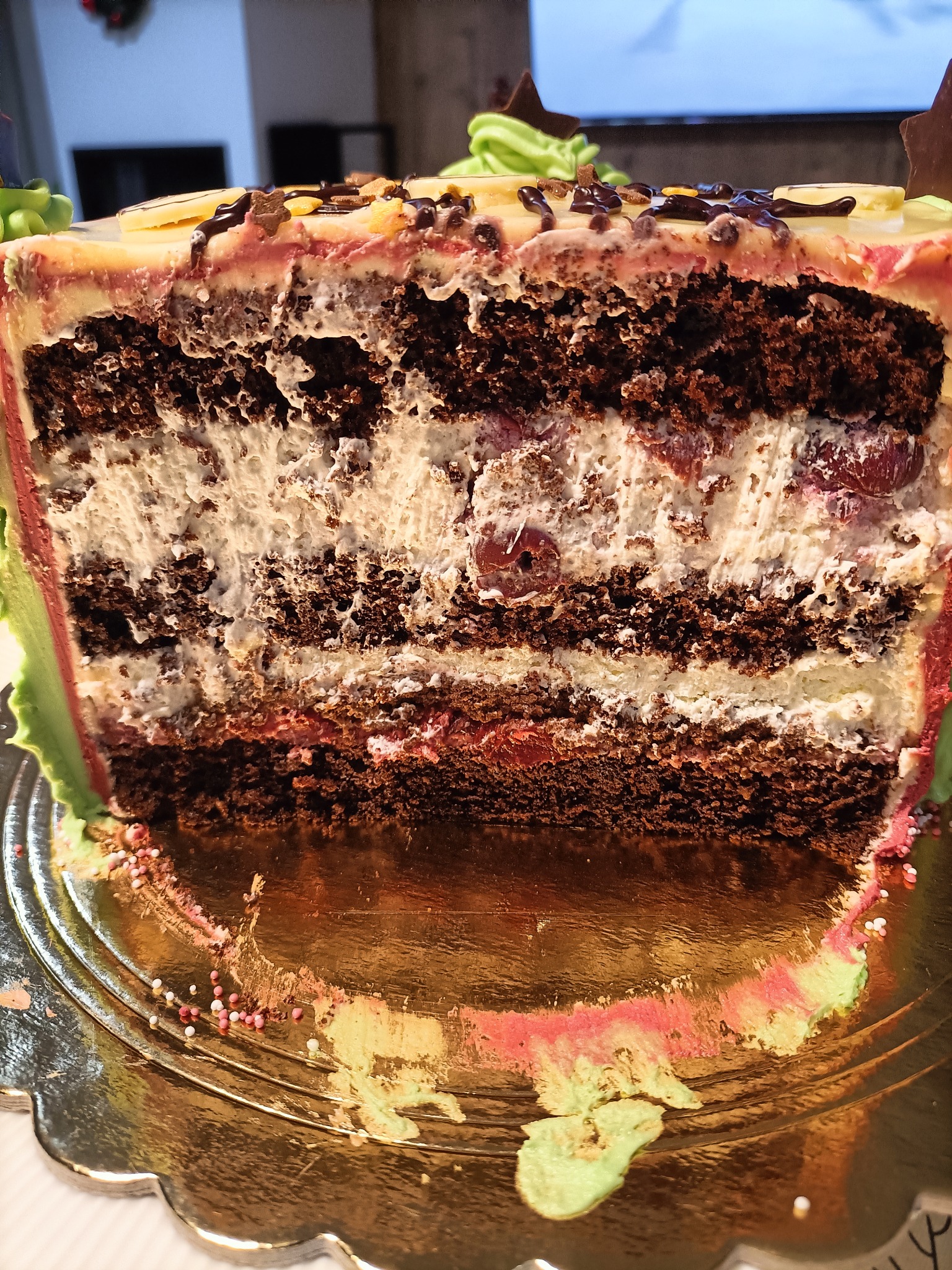 Przekrój tortu czekoladowego z kremem śmietankowym i wiśniami, udekorowany polewą i elementami z czekolady, na złotej paterze z rozsypanymi kolorowymi kuleczkami.