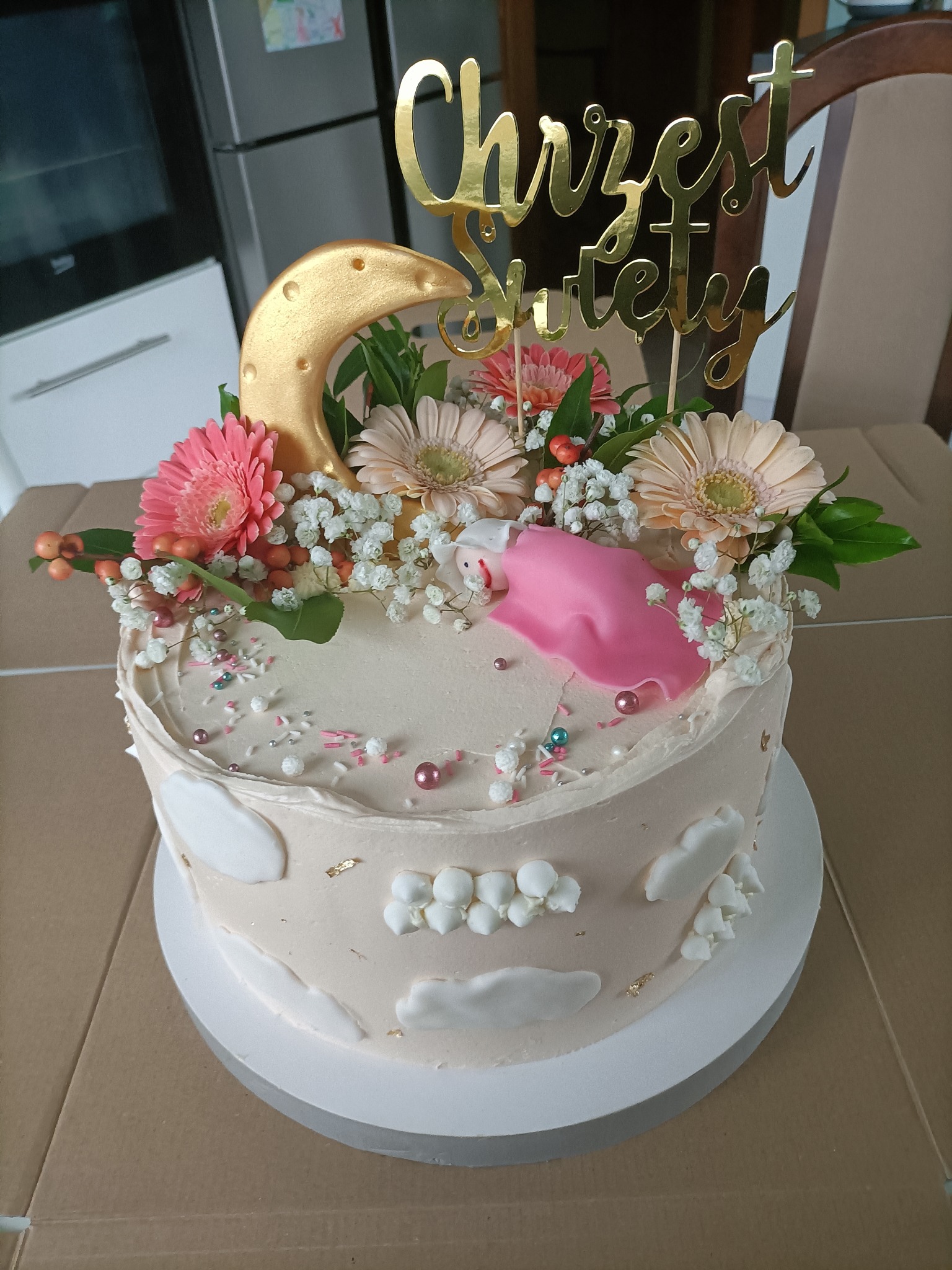 Tort kremowy z dekoracją z kwiatów, złotego księżyca i napisu 'Chrzest Święty', ozdobiony małymi chmurkami i złotymi płatkami.