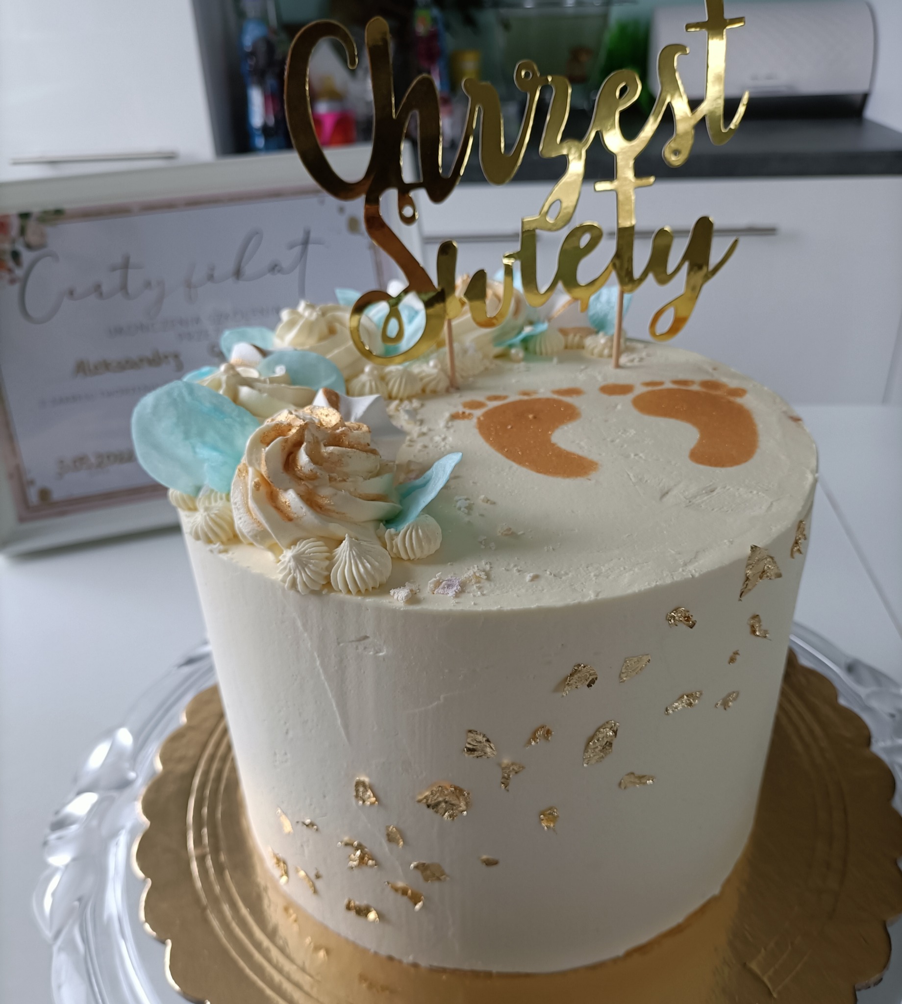 Tort na Chrzest Święty z białym kremem, złotymi płatkami, dekoracjami z kremu w kolorze ecru i błękitu oraz złotym napisem i odciskiem stóp dziecka.