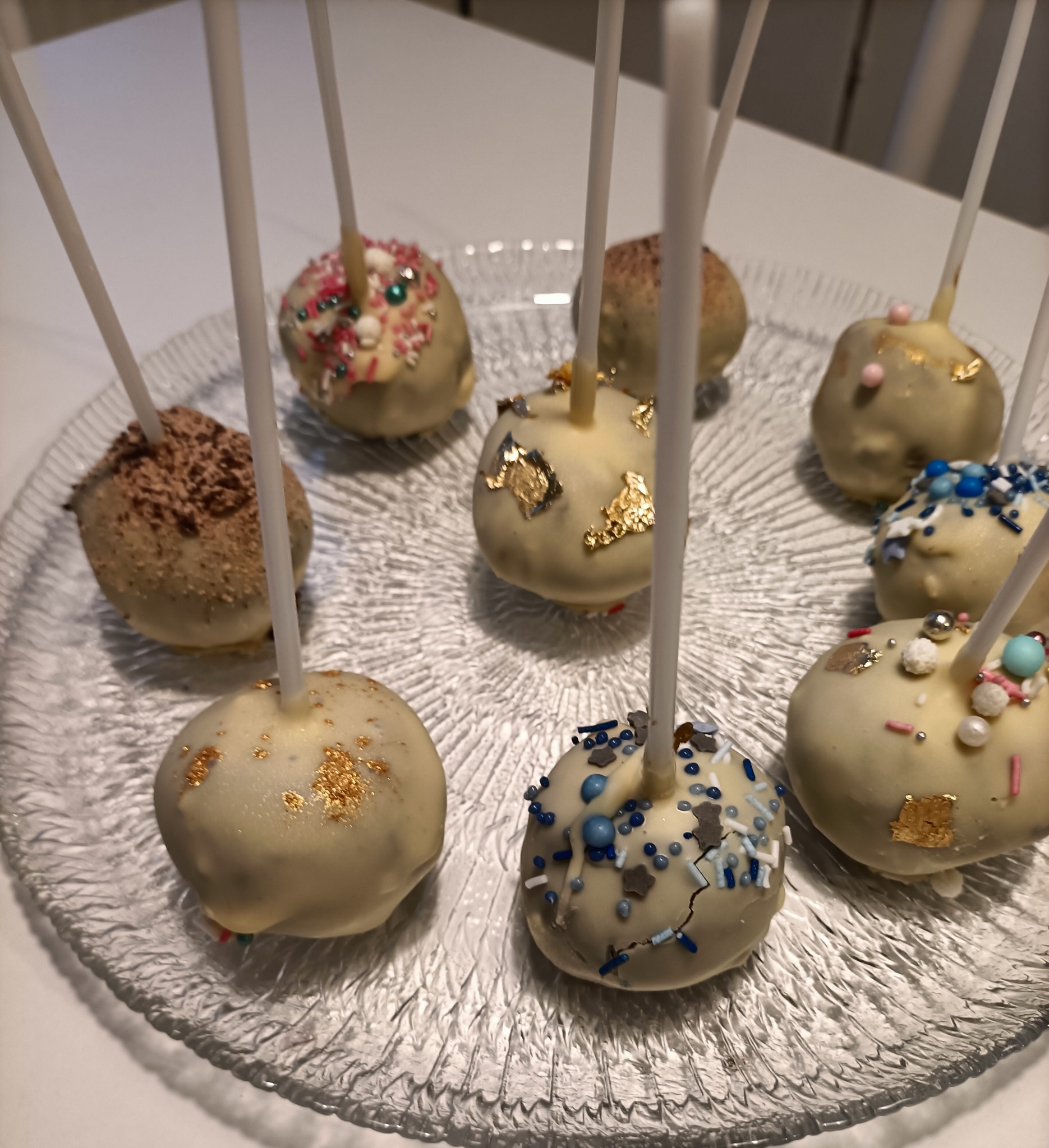 Kolekcja różnorodnych cake pops na szklanym talerzu: czekoladowe posypki, złote płatki, kolorowe kuleczki i gwiazdki.