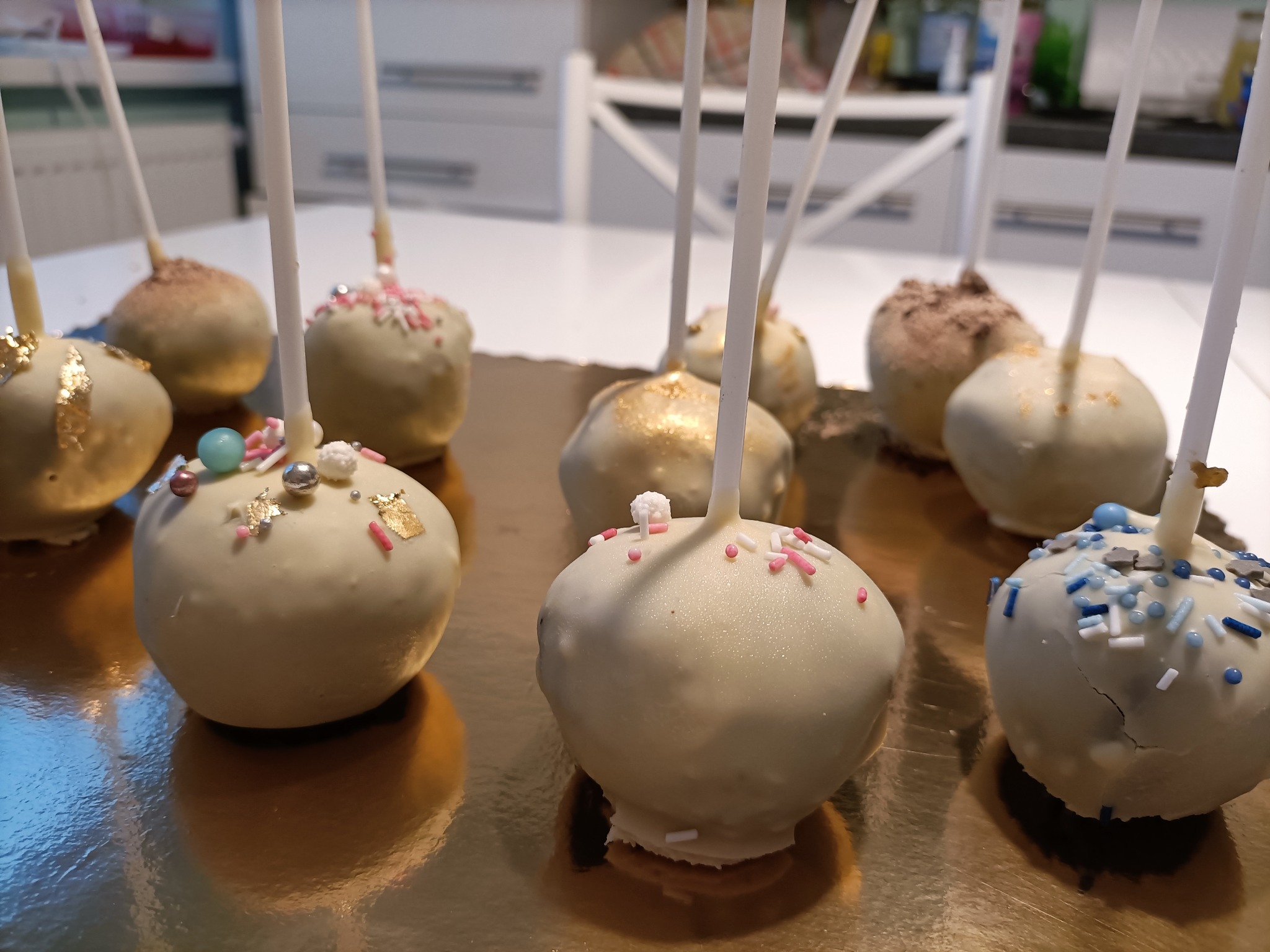 Różnorodna kolekcja cake pops z białej czekolady, udekorowanych kolorowymi posypkami, złotymi płatkami i niebieskimi koralikami, umieszczonych na złotej tacy.