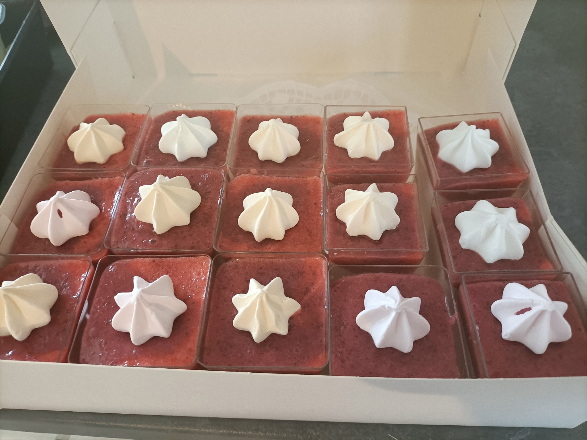 Kilkanaście porcji deseru truskawkowego w przezroczystych kwadratowych pojemnikach, udekorowanych kremowymi rozetkami w białym kartonowym pudełku.