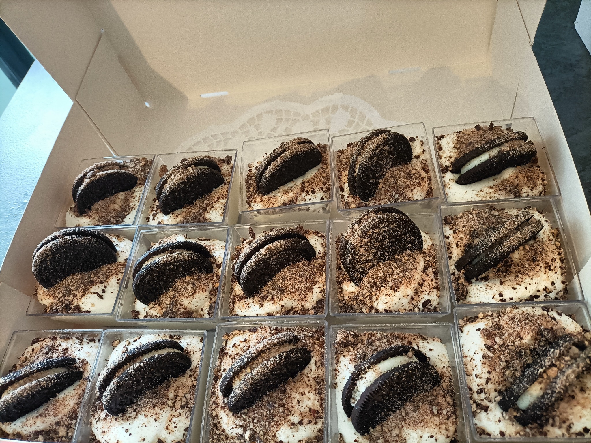 Karton z deserami w małych, przezroczystych pojemnikach: biała baza kremowa, posypka z pokruszonych ciastek czekoladowych i połówka ciastka Oreo na wierzchu.