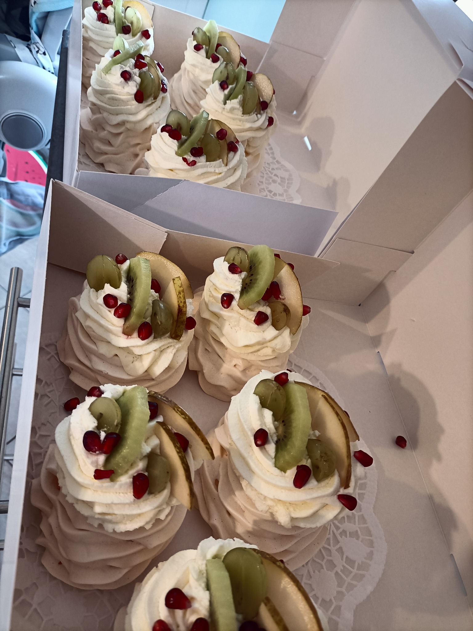 Bezy Pavlova udekorowane bitą śmietaną, kiwi, gruszką i granatem, ułożone w dwóch rzędach w białym kartonie na koronkowej serwetce.