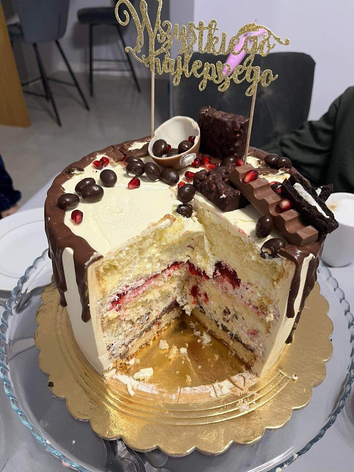 Tort z kremem, czekoladowymi polewami i dekoracjami, z wyciętym kawałkiem, ukazującym warstwy biszkoptu i owocowego nadzienia, umieszczony na szklanej paterze ze złotym podkładem. Na torcie napis...