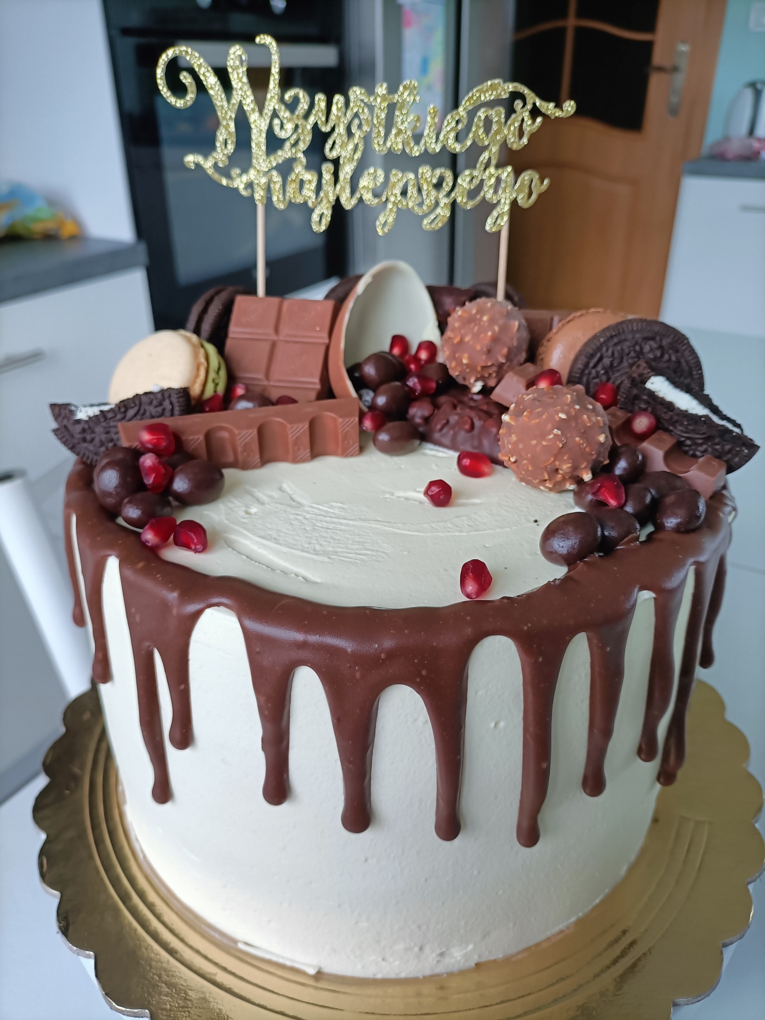 Tort urodzinowy z białym kremem, polewą czekoladową, dekoracjami z czekolady, ciastek Oreo, makaroników i granatu, zwieńczony złotym napisem 'Wszystkiego Najlepszego'.
