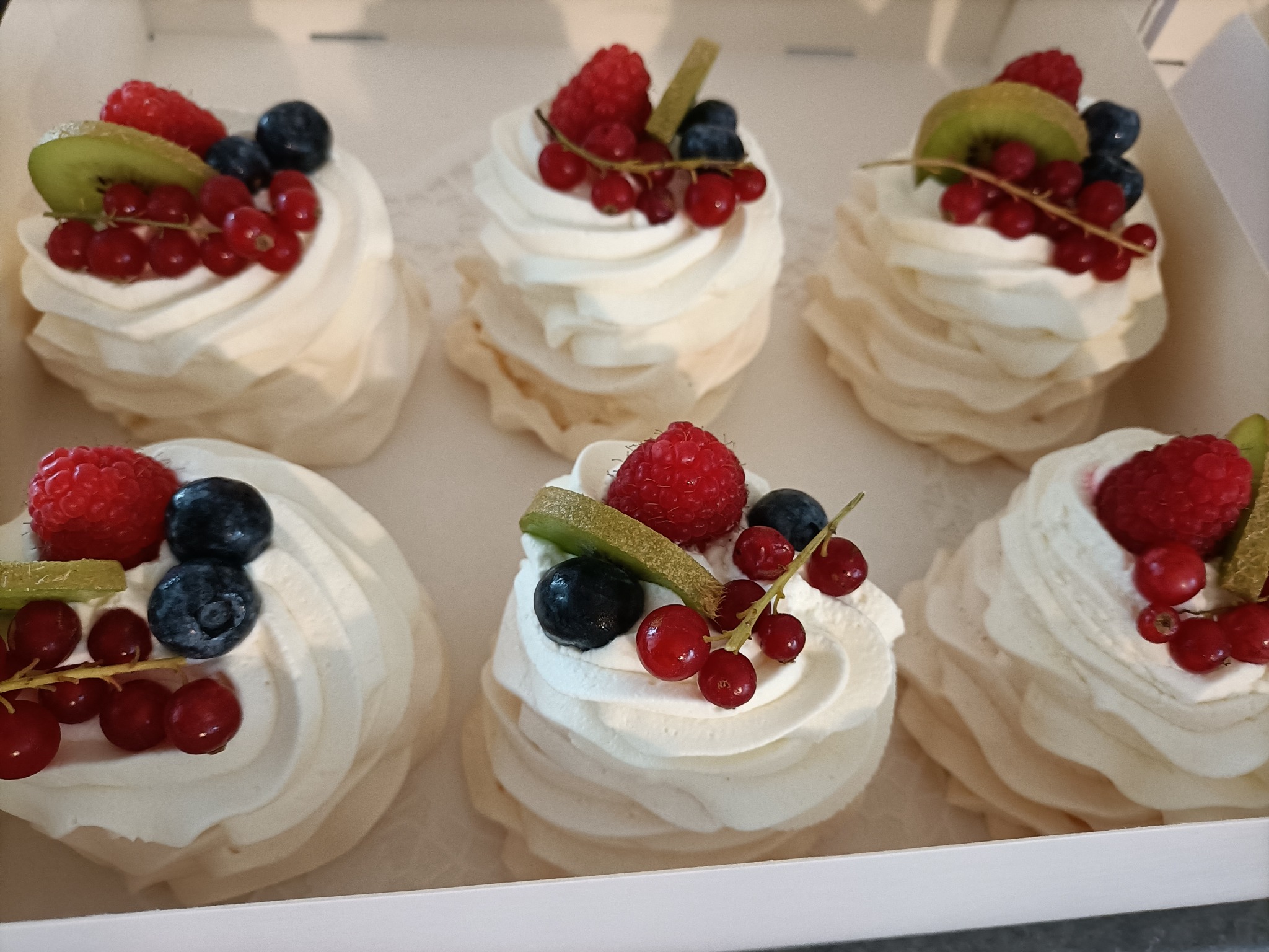 Sześć mini bez z bitą śmietaną, malinami, borówkami, plasterkami kiwi i gałązkami czerwonych porzeczek, ułożonych w białym pudełku.