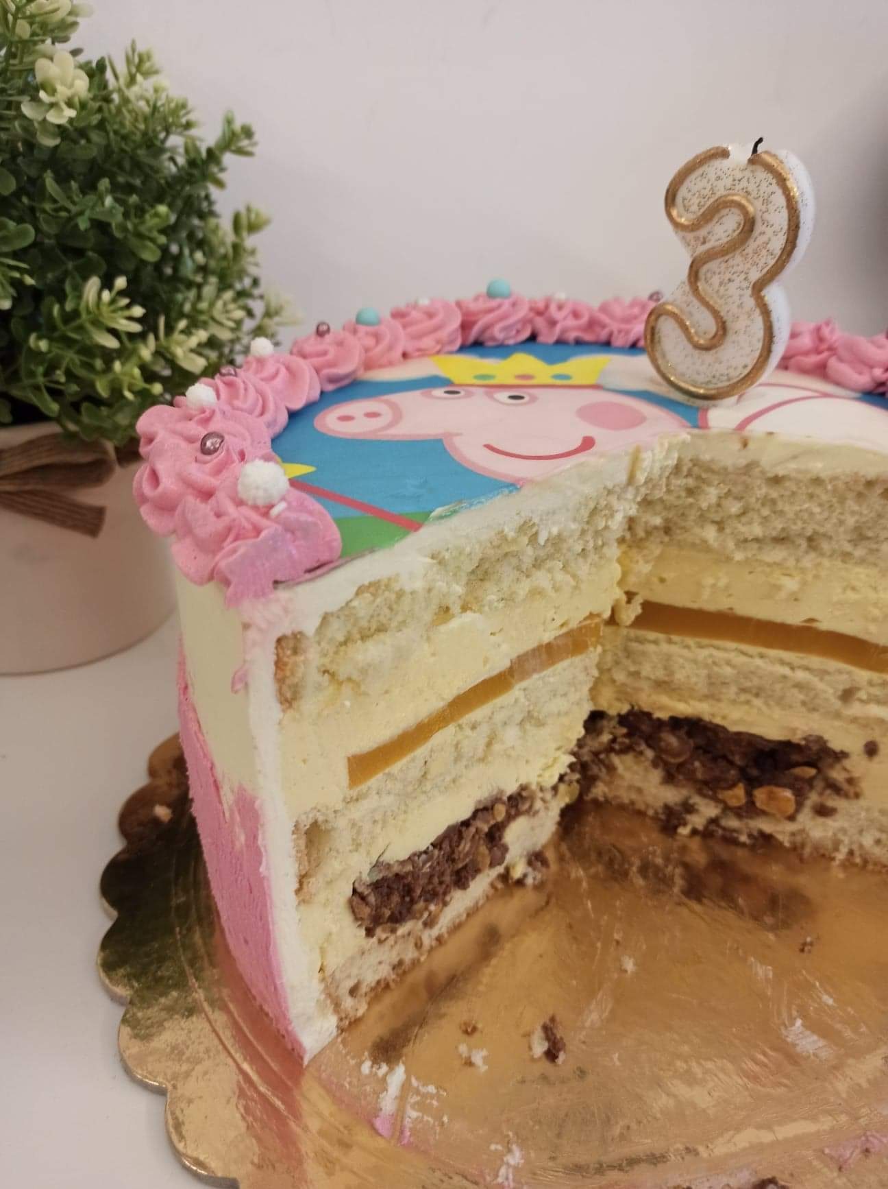 Przekrój tortu urodzinowego z wizerunkiem postaci z bajki, udekorowanego różowym kremem i świeczką w kształcie cyfry 3, obok doniczka z zieloną rośliną.