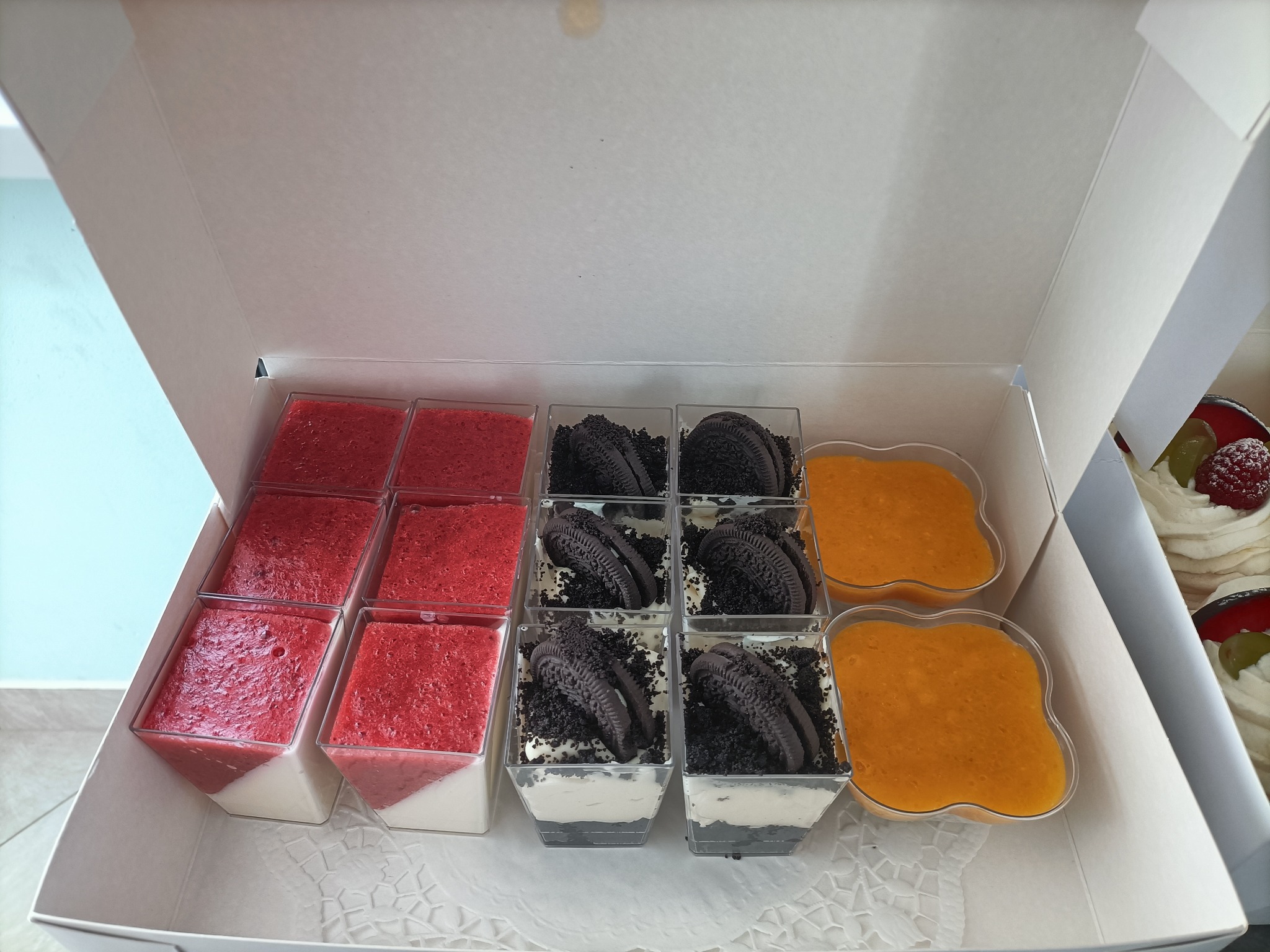 Zestaw deserów w pudełku: mus truskawkowy na śmietance, deser Oreo z kruszonką i kremem, oraz mus mango w ozdobnych pojemnikach.