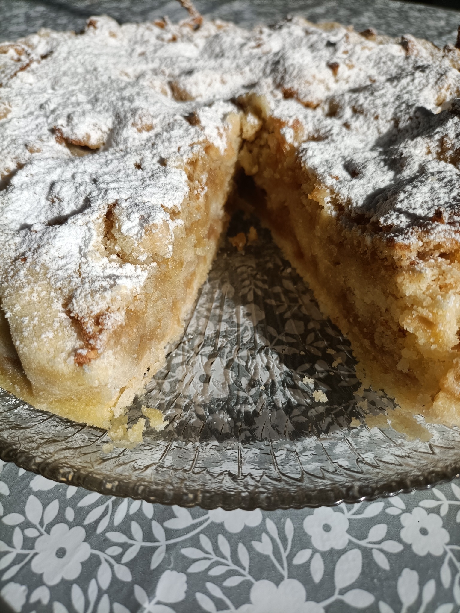 Jabłecznik posypany cukrem pudrem, z wyciętym kawałkiem, na szklanym talerzu z wzorem liści na stole.