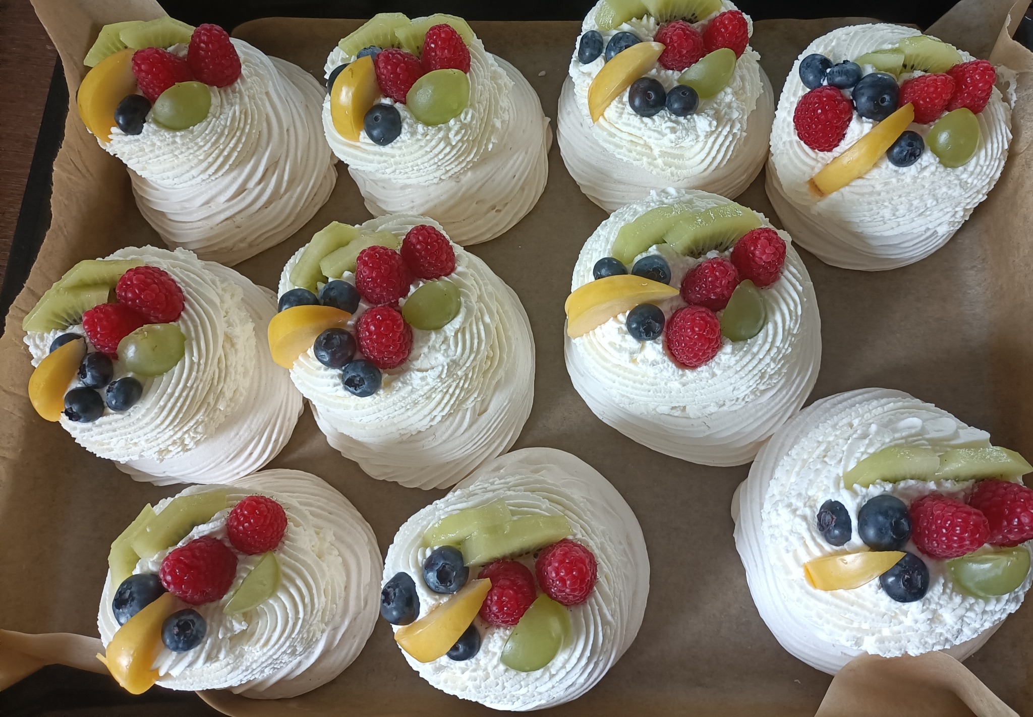 Rząd bezowych babeczek z bitą śmietaną i świeżymi owocami: malinami, borówkami, kiwi, winogronami i brzoskwiniami, ułożonych na papierze do pieczenia.