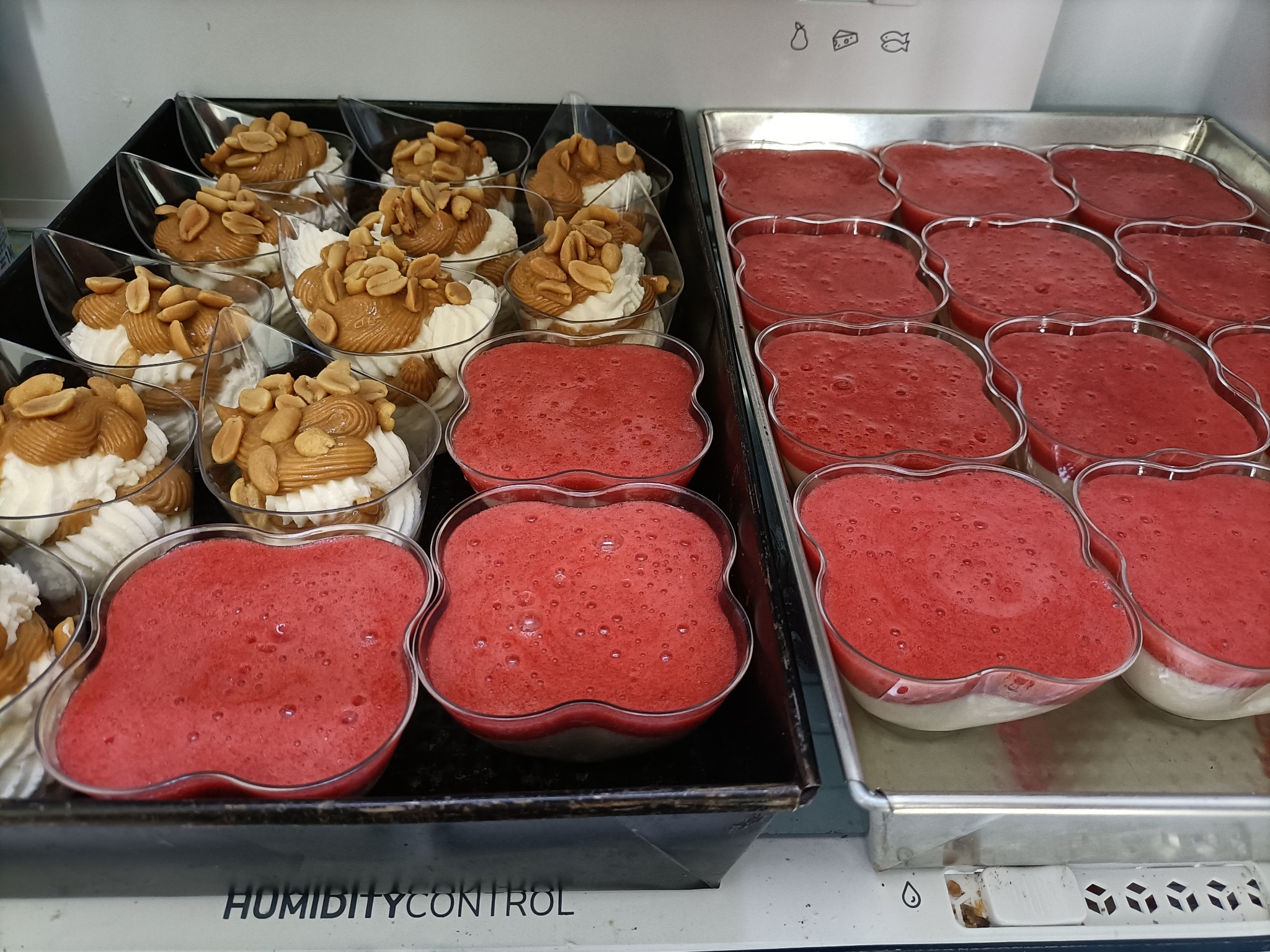 Dwa rodzaje deserów w plastikowych pojemnikach: kremowe desery z orzechami i czerwone musy owocowe na tacy.