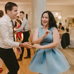 Para tańczy na parkiecie podczas wesela, kobieta w błękitnej sukience śmieje się, mężczyzna w białej koszuli i czarnych spodniach trzyma ją za ręce.