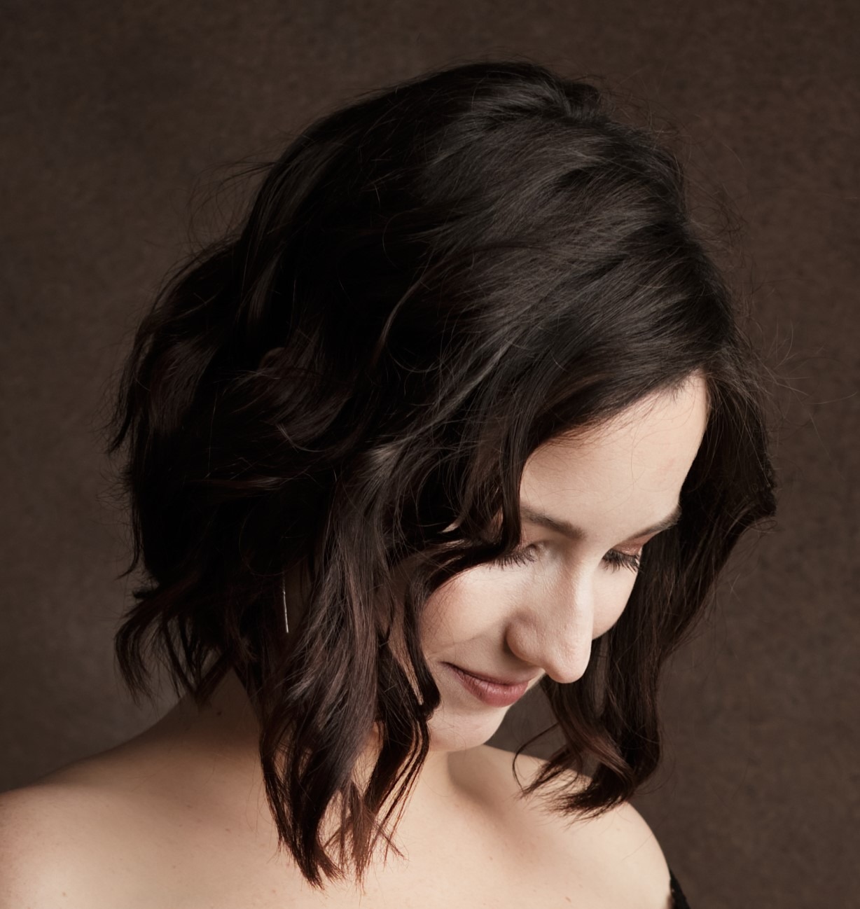 Portret kobiety z ciemnymi, falowanymi włosami, spoglądającej w dół na brązowym tle.