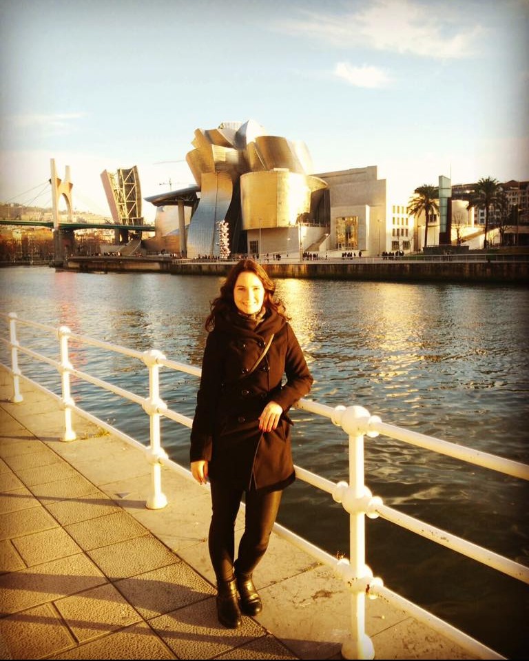 Kobieta w czarnym płaszczu pozuje na tle Muzeum Guggenheima w Bilbao, odbijającego się w wodach rzeki Nervión.