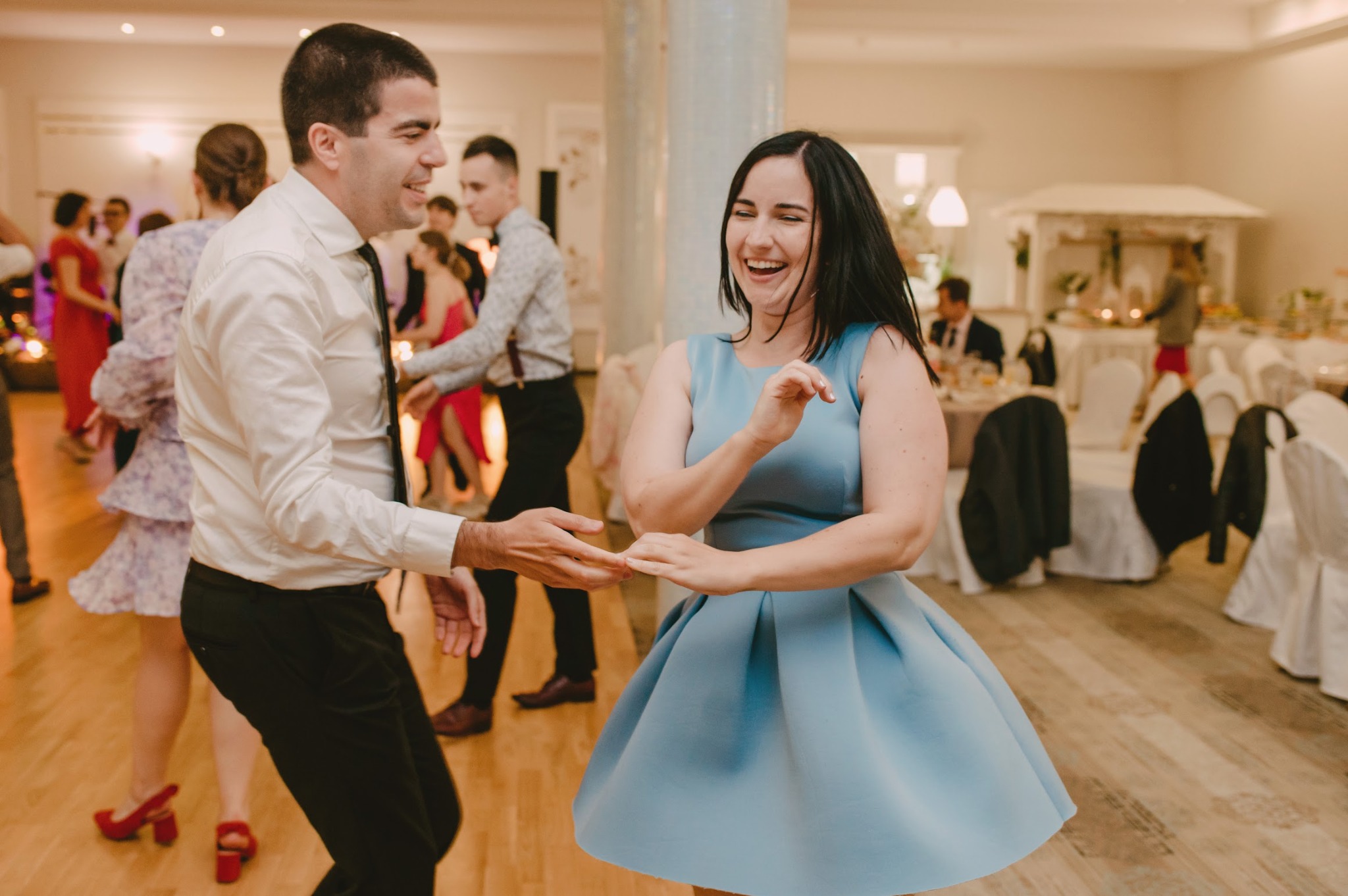 Para tańczy na parkiecie podczas wesela, kobieta w błękitnej sukience śmieje się, mężczyzna w białej koszuli i czarnych spodniach trzyma ją za ręce.