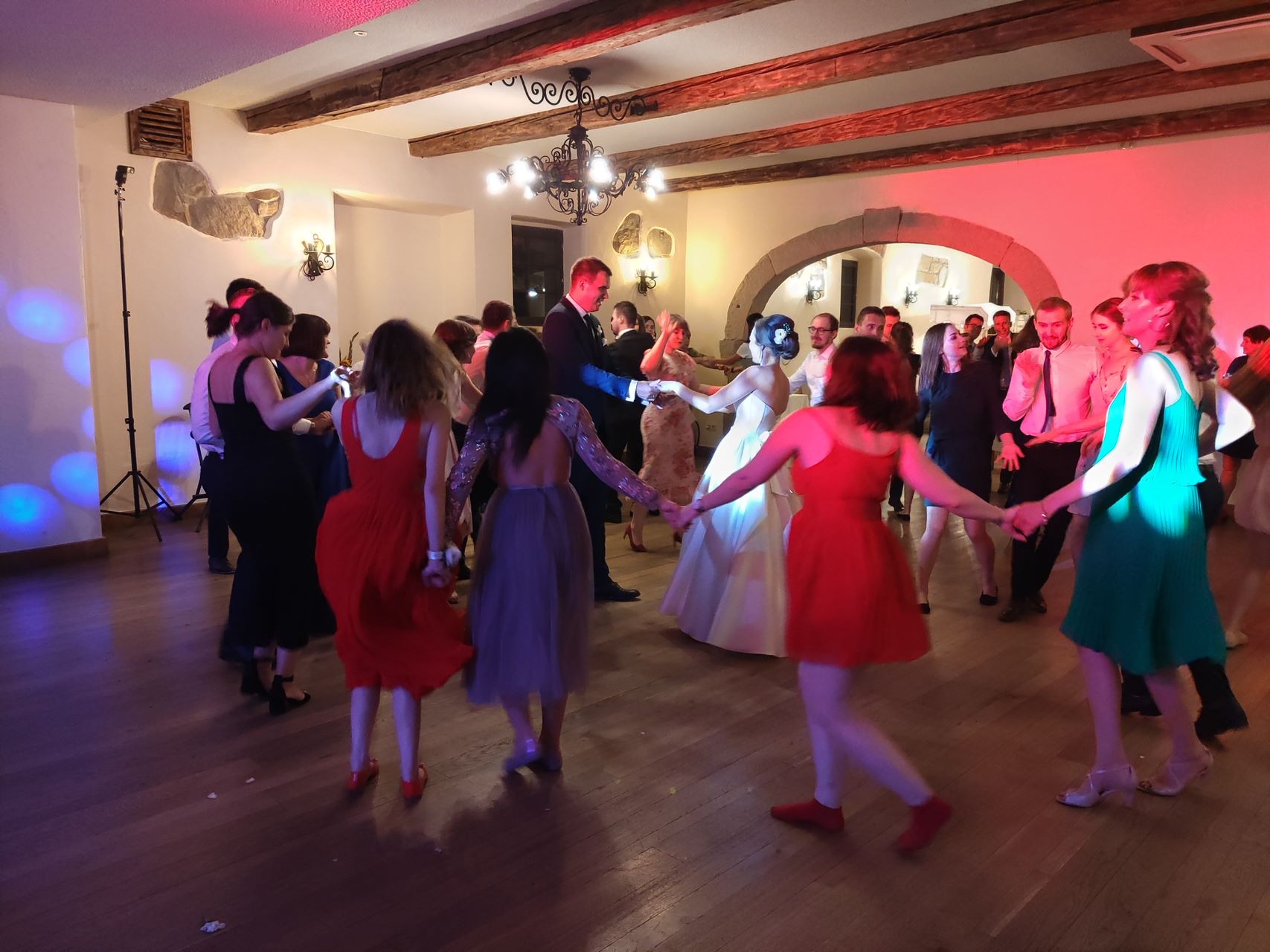 Grupa ludzi w sukienkach i garniturach tańczy w kole podczas wesela w sali z drewnianym sufitem i kamiennymi elementami dekoracyjnymi.