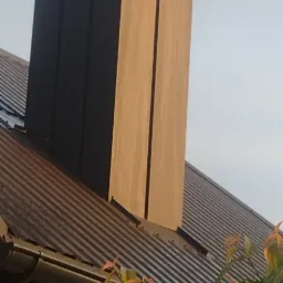 Nowoczesny komin obłożony panelami w dwóch odcieniach na dachu z blachy trapezowej, widok z dołu przez gałęzie drzewa.