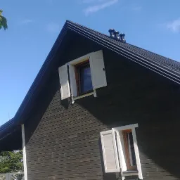 Dom z ciemnego drewna z grafitowym dachem, białymi okiennicami i rynnami, widok z dołu w słoneczny dzień.