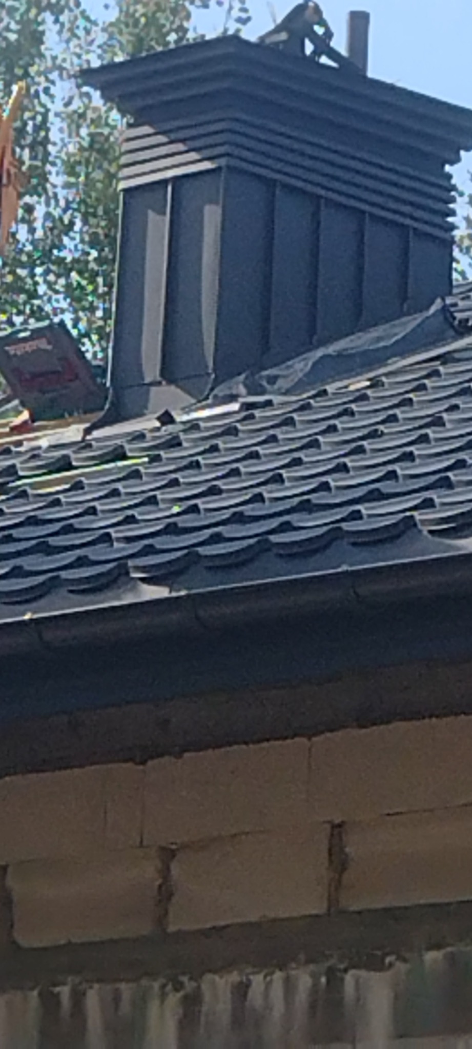 Czarny komin z obróbkami blacharskimi na dachu z blachodachówki, widoczny fragment ściany budynku z pustaków.