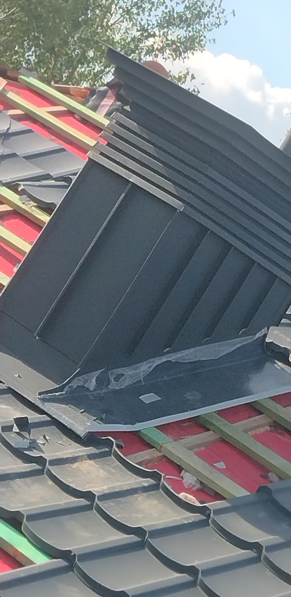 Montaż obróbki blacharskiej na dachu z czarnej dachówki, widoczne elementy konstrukcji dachu i folia ochronna.