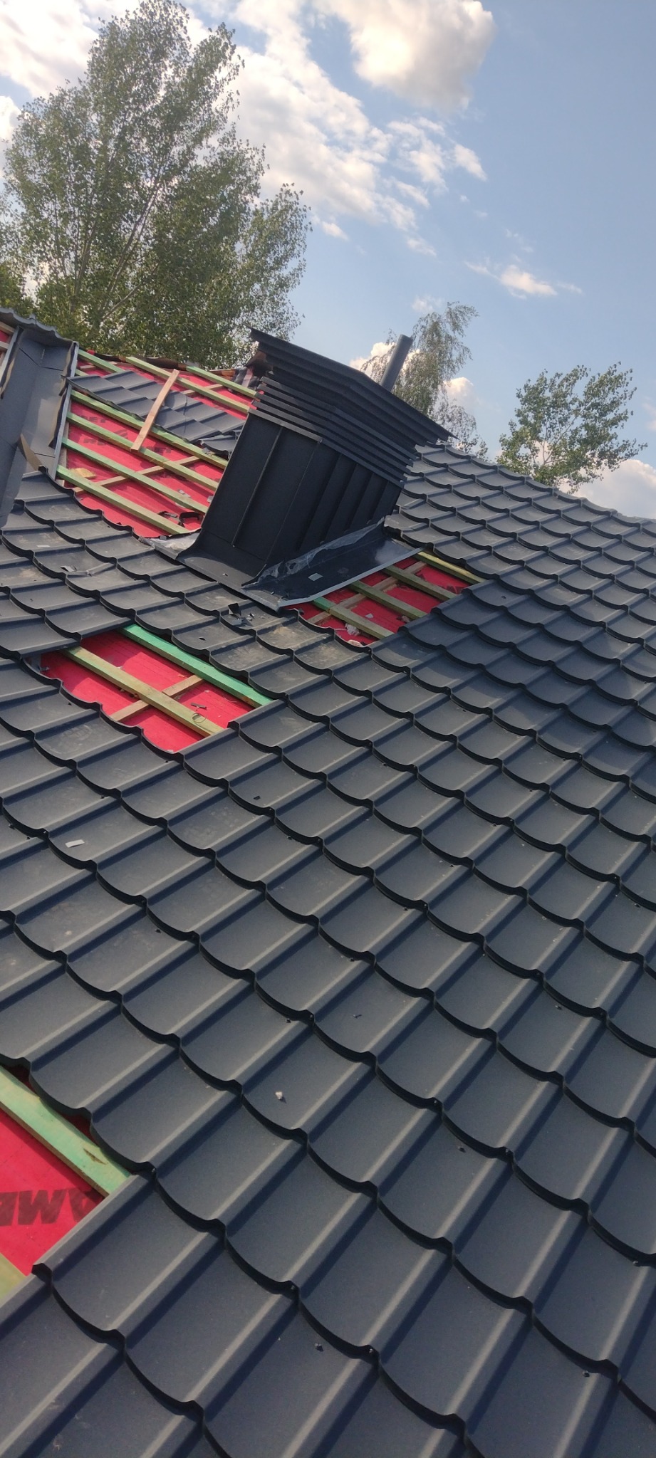 Częściowo położony dach z czarnej blachodachówki, widoczne fragmenty czerwonej membrany i drewnianej konstrukcji, komin wentylacyjny, tło: błękitne niebo z chmurami i korony drzew.