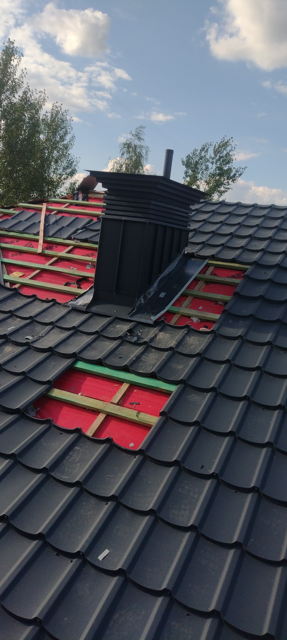Częściowo zdjęte czarne dachówki, widoczna czerwona membrana i drewniana konstrukcja dachu oraz komin z blachy na tle błękitnego nieba.