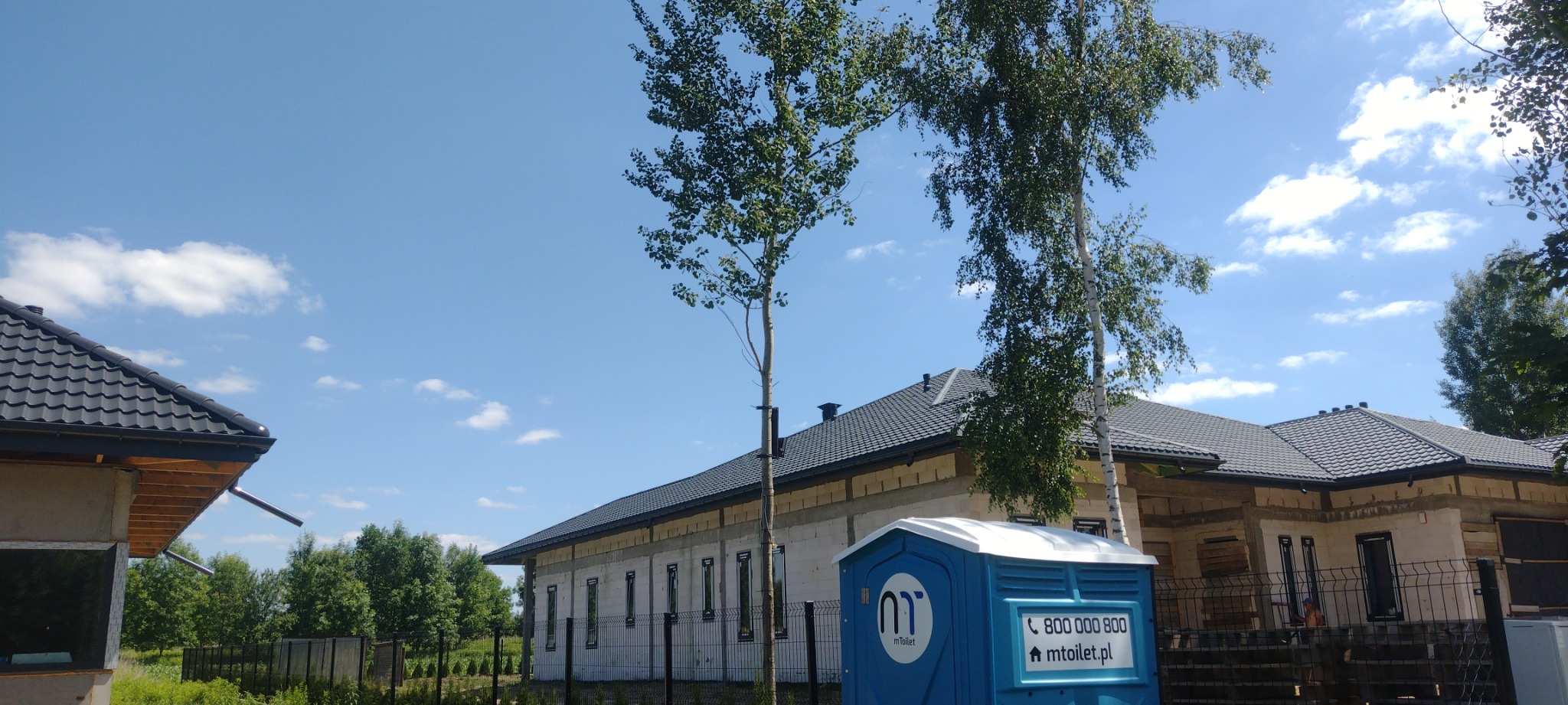 Nowo położony, grafitowy dach z blachodachówki na budynku w stanie surowym w Starej Kornicy, z widoczną toaletą przenośną na pierwszym planie i młodymi brzozami.