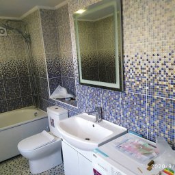 FORTUNA - Ukończona łazienka w Poznaniu z mozaikową glazurą w odcieniach błękitu i beżu, z podświetlanym lustrem, białą armaturą i pralką LG w rogu. Widoczny fragment podłogi wyłożonej pasującymi płytkami.