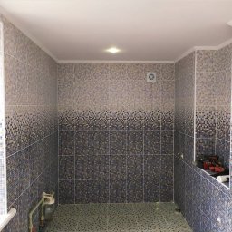 FORTUNA - Wykończona łazienka z mozaikową glazurą w odcieniach beżu i granatu, widoczna wentylacja i rury doprowadzające wodę.