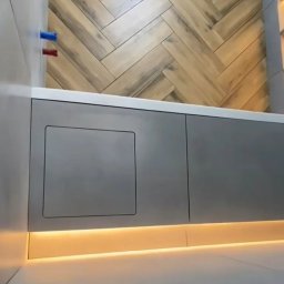 FORTUNA - Szara zabudowa pod wanną z ukrytymi drzwiczkami rewizyjnymi, podświetlona listwą LED, na ścianie płytki imitujące drewno ułożone w jodełkę, widoczne rury instalacji wodnej.