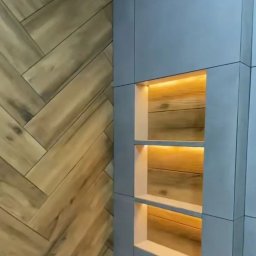 FORTUNA - Ściana z drewnianą okładziną ułożoną w jodełkę, sąsiadująca z szarą zabudową z trzema podświetlanymi półkami, ukazującymi drewniane deski wewnątrz.