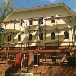 FORTUNA - Dwóch pracowników na rusztowaniu podczas prac na elewacji budynku z ceglaną dobudówką, widoczne okna z łukowym wykończeniem i błękitne niebo.